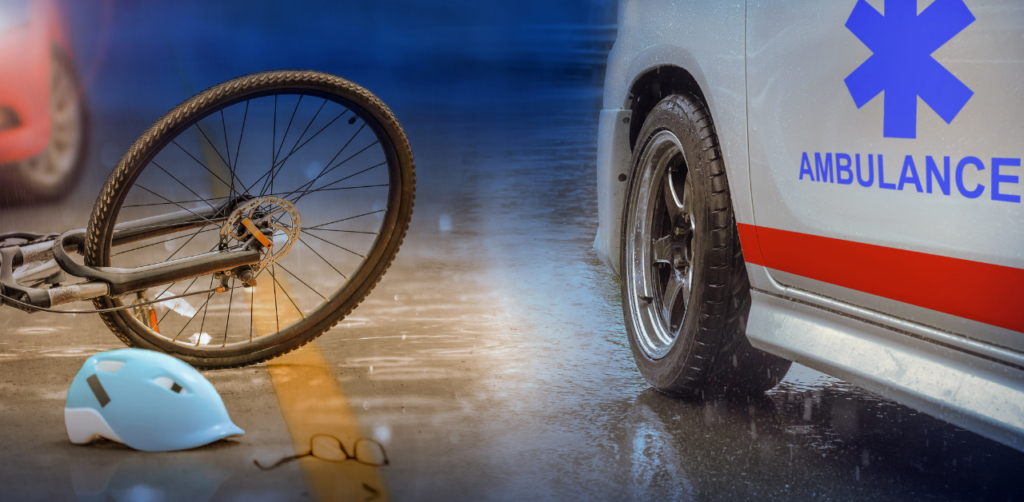 Fallen bicycle, blue helmet, and glasses on a wet road beside an ambulance, indicating a recent accident in rainy weather