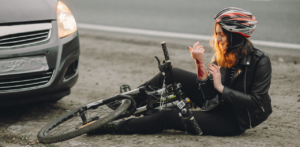 Woman wearing a helmet and black jacket sits on the roadside next to a bicycle, examining a scrape on her arm, with a car nearby