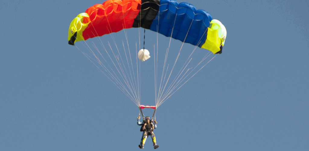 A skydiver glides through the air with a vibrant rainbow-colored parachute against a clear blue sky