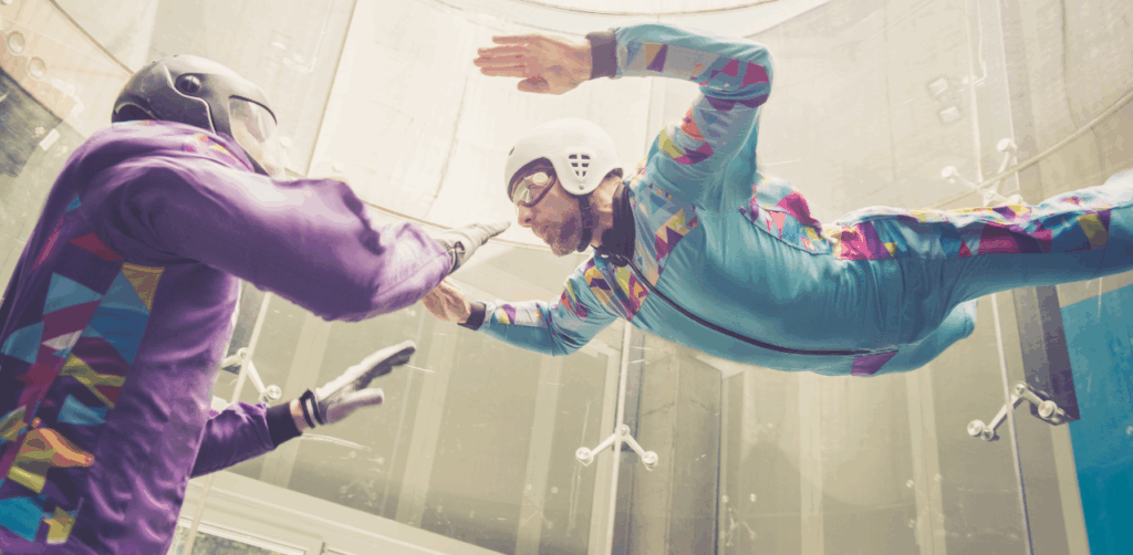 Two men practice body flight in an indoor skydiving wind tunnel wearing colorful jumpsuits and helmets