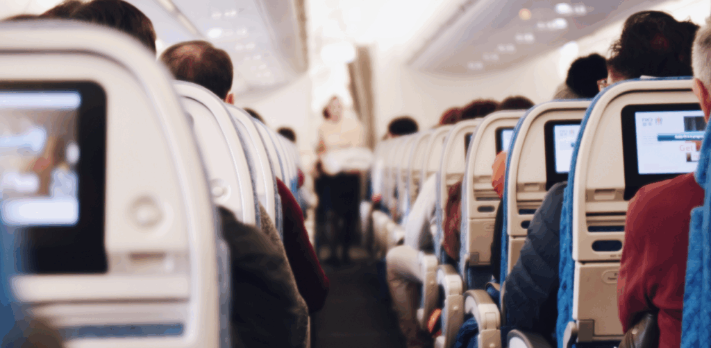 Interior view of a crowded airplane cabin, showing passengers seated with personal screens, illustrating compliance with air travel safety protocols