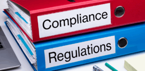 Two large binders labeled Compliance and Regulations are stacked on a desk, symbolizing legal and policy documentation in a corporate or regulatory setting
