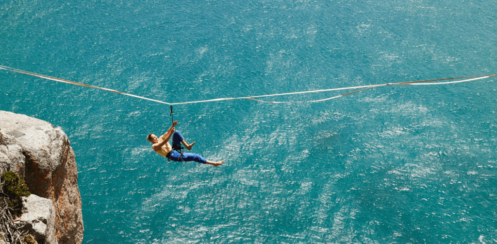 Person in a harness glides across a zip line high above the ocean between two cliffs, capturing the thrill and risk of high-altitude outdoor recreation