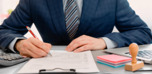 Businessperson in a suit signing a document at a desk, with office supplies and a calculator nearby