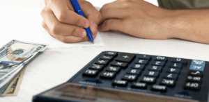 Person writes on paper next to a stack of hundred-dollar bills and a calculator, symbolizing financial planning or evaluating a personal injury settlement amount