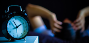A close-up of an alarm clock showing 5:50 AM, with a blurred figure holding their head in the background, suggesting stress or insomnia