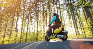 A person rides a yellow scooter on a forest road, with sunlight filtering through tall trees in the background