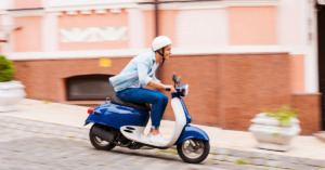 A person wearing a white helmet rides a blue scooter on a cobblestone street, passing a peach-colored building