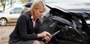 A woman kneeling by a damaged car, using a tablet, likely documenting or assessing the damage from an accident