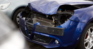 The front end of a blue car is severely damaged, with the bumper and radiator exposed, indicating the aftermath of a collision