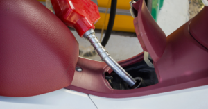 Close-up of a red fuel pump nozzle inserted into the gas tank of a maroon scooter seat at a gas station