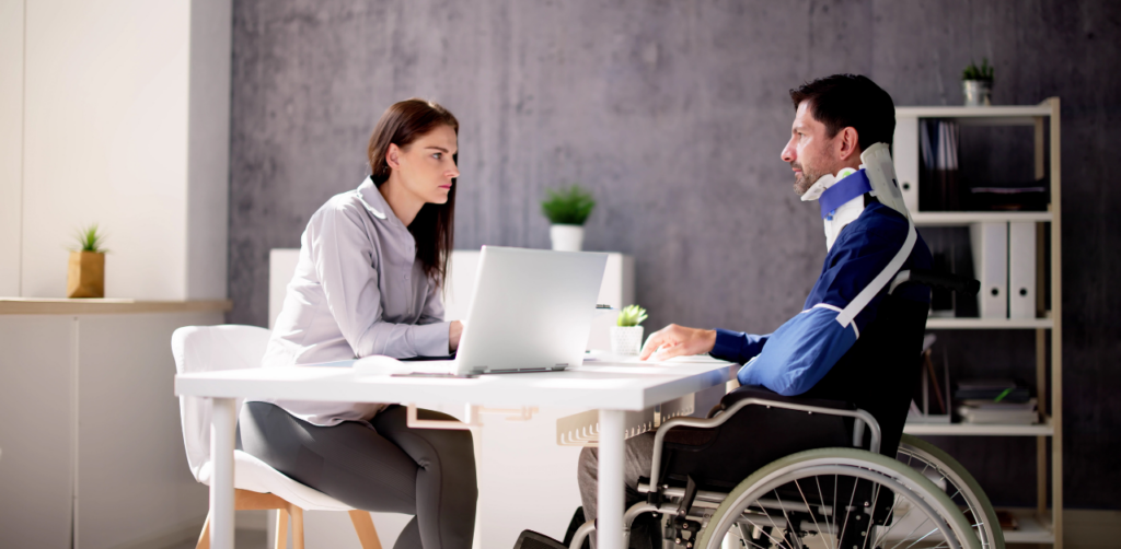 A man in a wheelchair with a neck brace talking to a woman at a desk with a laptop in an office setting