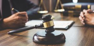 A gavel rests on a wooden table with two people in a discussion in the background, one holding a pen