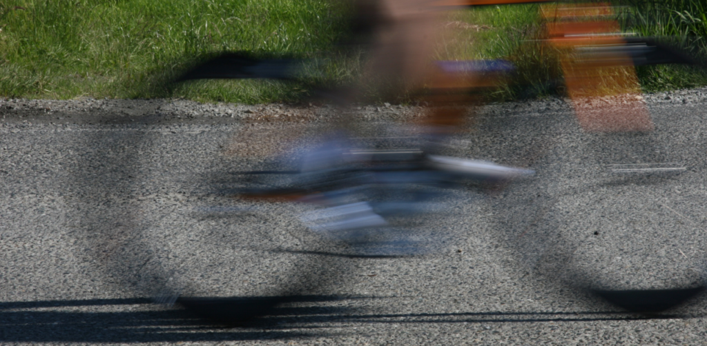 Blurry motion capture of a cyclist speeding by on a paved road, with green grass visible at the edge of the image