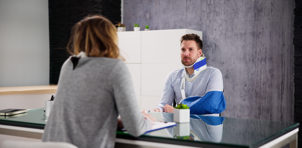Man with a neck brace and arm in a sling sits across a desk from a woman taking notes, in a modern office setting