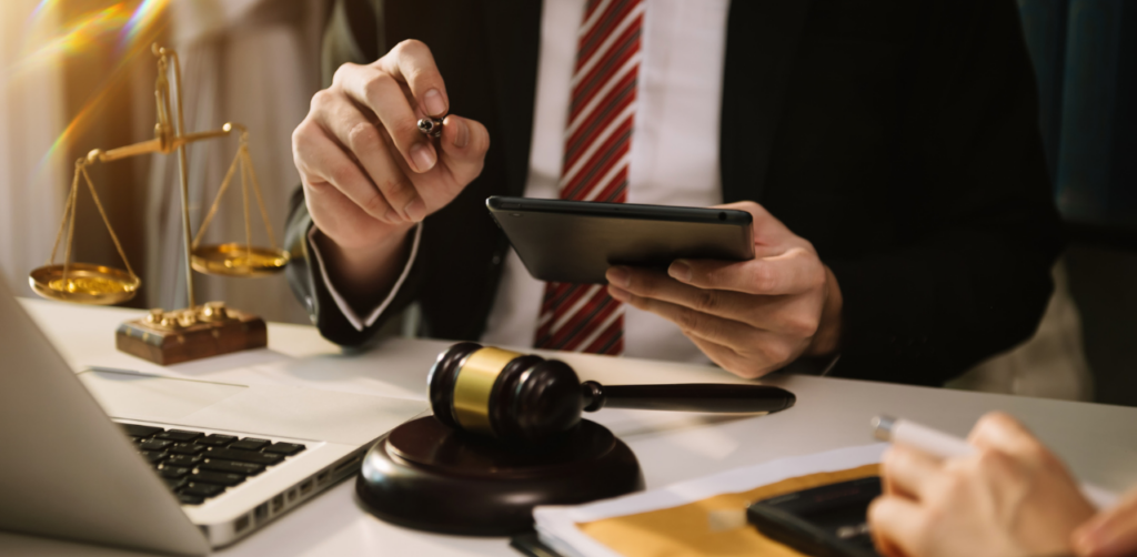 Person in a suit holding a tablet, with a gavel and scales of justice on the desk, symbolizing a legal or courtroom setting