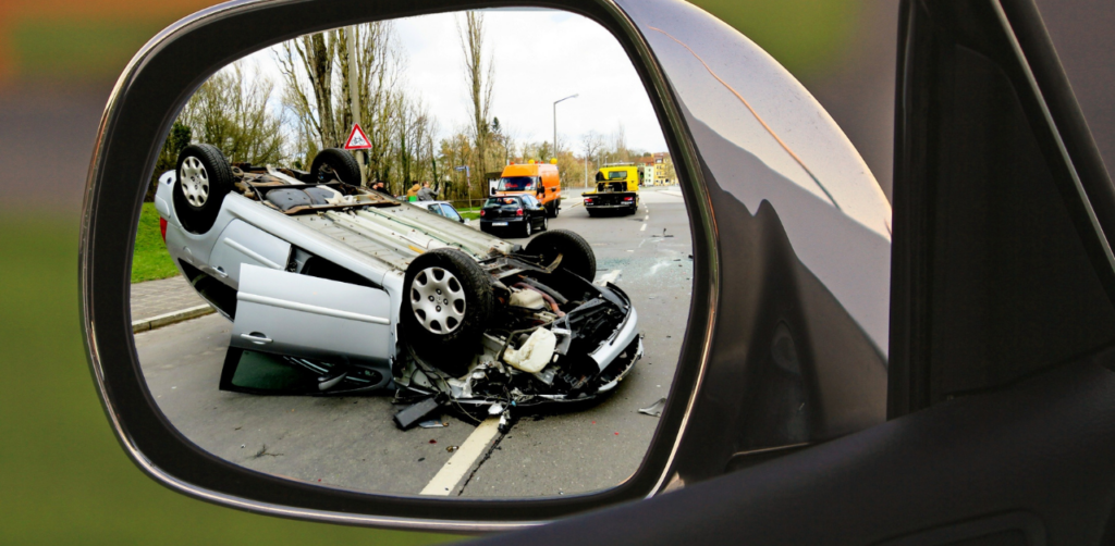 Rearview mirror showing an overturned car in the road, with emergency vehicles and people in the background attending the scene