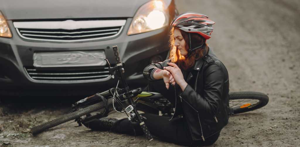 Woman in a helmet and leather jacket sitting on the ground next to a bicycle, inspecting her wrist in front of a car with headlights on