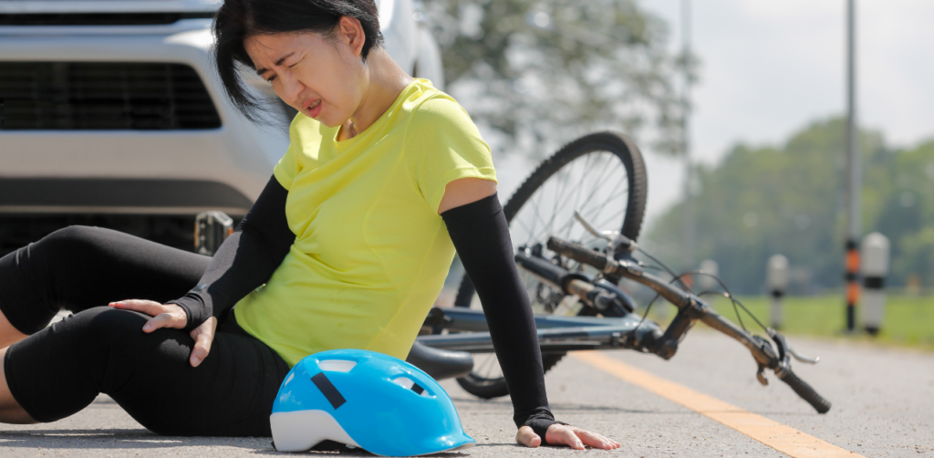 Woman in a yellow shirt sitting on the road next to a bicycle, grimacing in pain and holding her knee, with a blue helmet nearby and a car in the background
