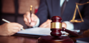 A wooden gavel in the foreground, with two people in suits writing on legal documents in the background