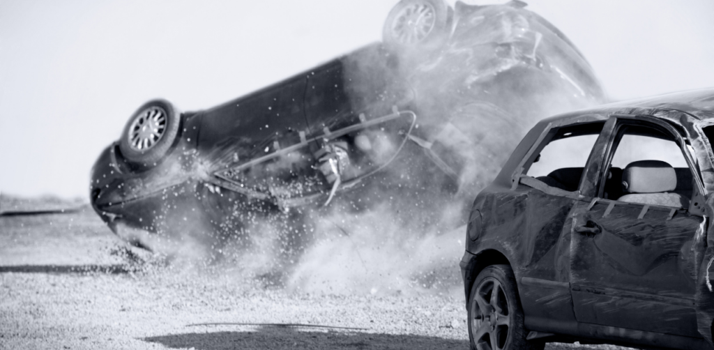 Black-and-white scene of a car crash with one vehicle overturned and another damaged in the foreground