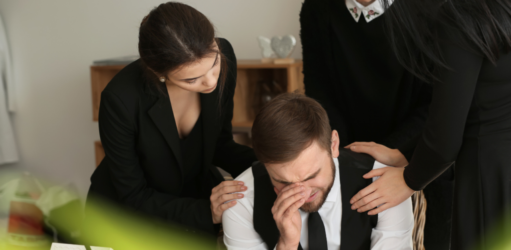 Grieving man in a suit being comforted by others, with hands on his shoulders, suggesting a mourning or support scene