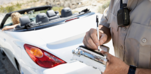 Police officer writes a traffic ticket beside a stopped white convertible on a sunlit roadside