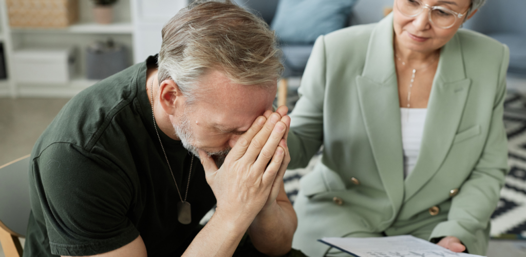 A distressed man with gray hair, seated and covering his face, comforted by an older woman holding a notepad