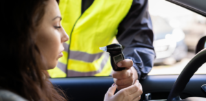 A driver in a car blowing into a breathalyzer held by a police officer wearing a reflective vest