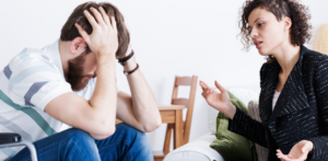 A woman with curly hair talking to a distressed man seated with his head in his hands, in a casual indoor setting