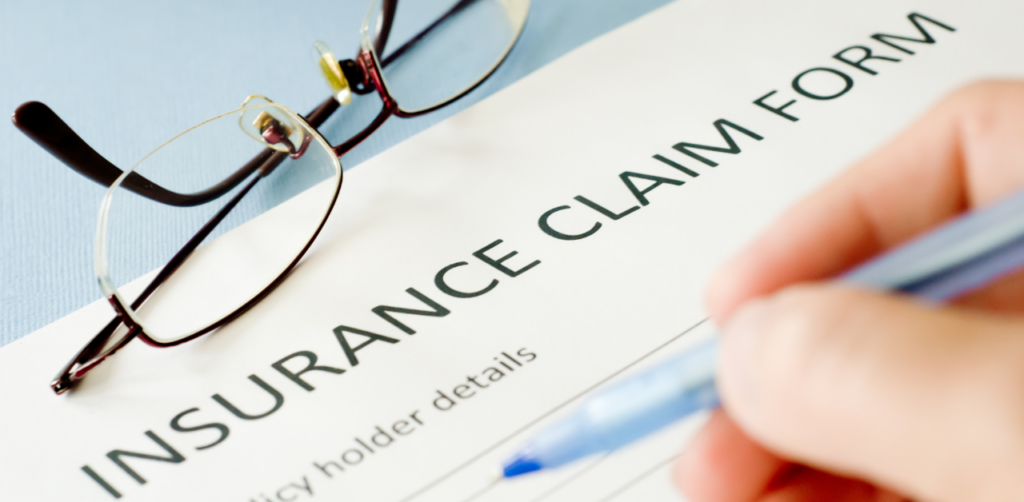 Close-up of a hand holding a pen over an insurance claim form, with reading glasses placed on top