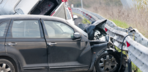 A black car with severe front-end damage is crashed into a highway guardrail, with debris and emergency vehicles in the background