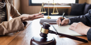 Close-up of a legal meeting with a gavel on the desk; one person gestures while another writes, with scales of justice visible in the background