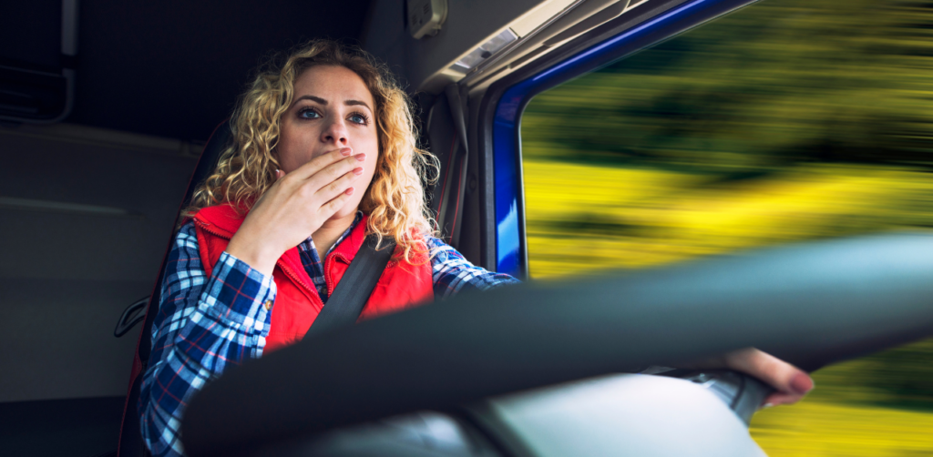 Concerned woman driving a vehicle covers her mouth with one hand, with blurred greenery visible through the window