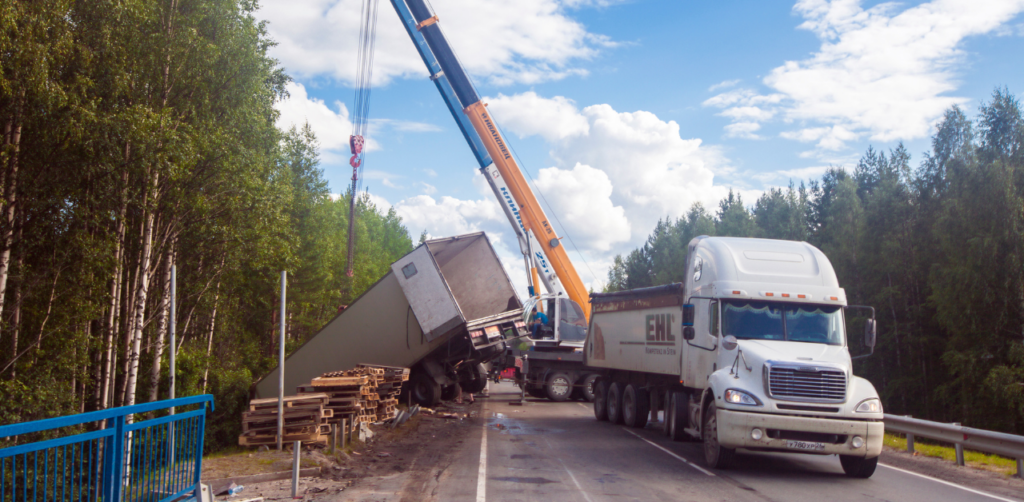 Crane lifts a tipped-over trailer on a road lined with trees, while a white truck waits nearby. Logs are scattered near the accident site