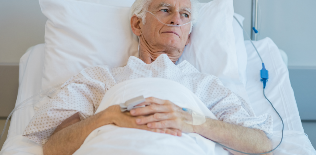 Elderly man in a hospital bed with an oxygen tube and IV, looking contemplative