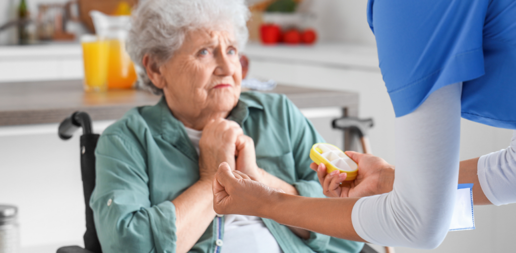 Elderly woman in a wheelchair looking hesitant as a caregiver offers her a pill organizer