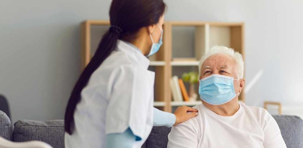 Healthcare worker wearing a mask comforting an elderly man in a mask, sitting on a couch