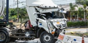 Heavily damaged white truck with a crumpled cab is parked on a roadside near a restaurant and greenery