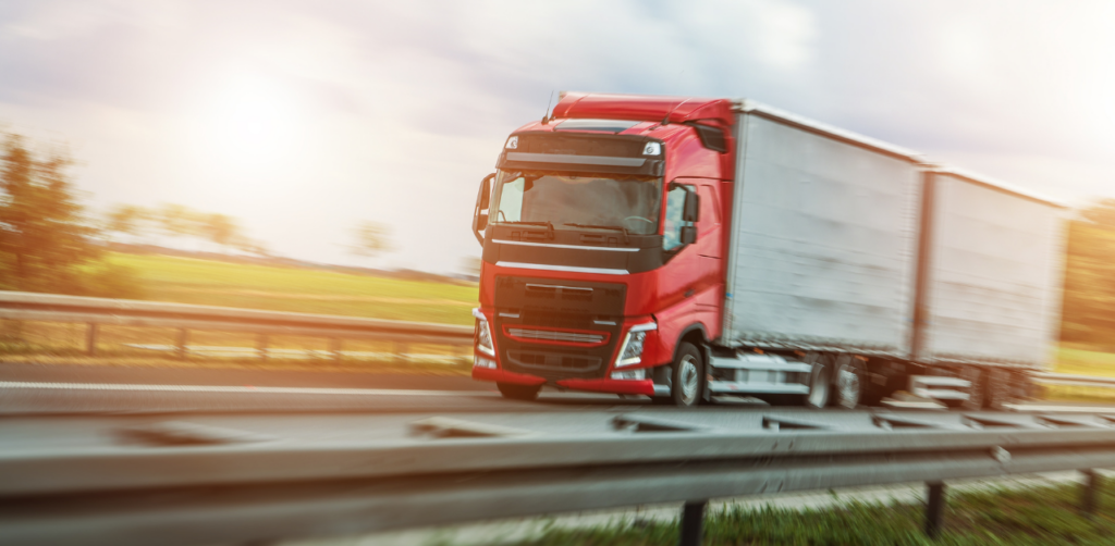 Red semi-truck with a trailer drives along a highway, passing green fields under a bright sky