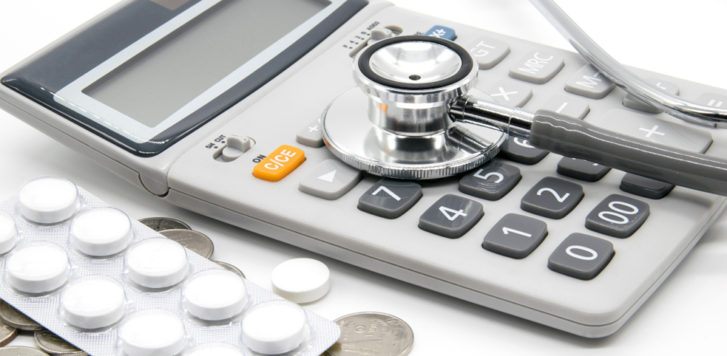 Stethoscope rests on a calculator, surrounded by coins and a blister pack of pills, symbolizing healthcare costs