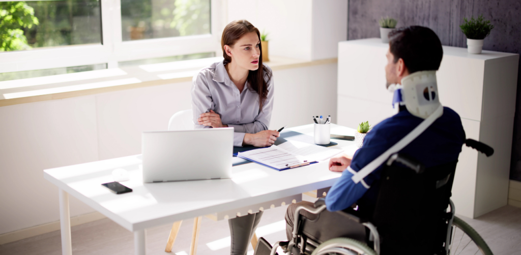 Woman interviews a man in a neck brace and wheelchair in a bright office, with a laptop and paperwork on the desk