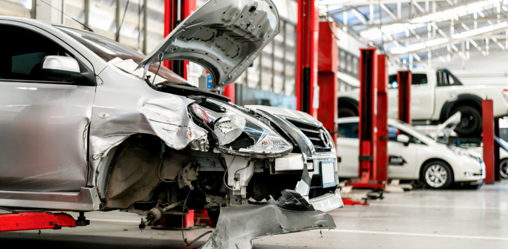 Damaged silver car with an open hood in an auto repair shop, surrounded by car lifts and other vehicles