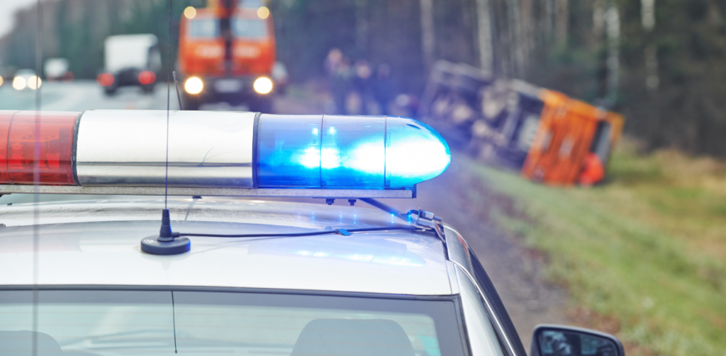Police car with flashing blue lights at a roadside accident involving an overturned truck in the background