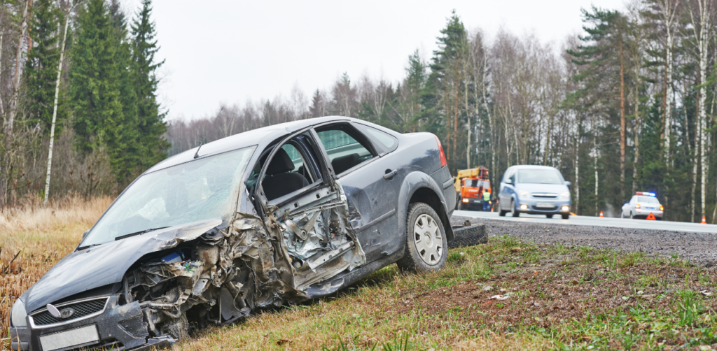 Heavily damaged dark-colored car sits off-road after an accident, with police and tow trucks nearby