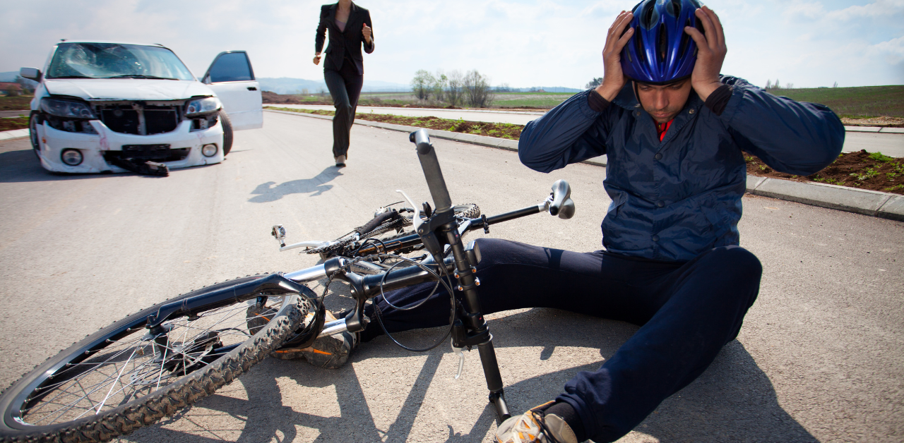 Cyclist sitting on the road holding his head after an accident with a damaged car and a bent bicycle