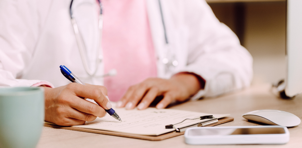 Doctor writing notes on a clipboard at a desk, with a stethoscope around their neck and medical tools nearby