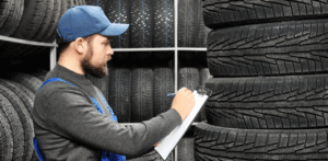 Mechanic in uniform and cap inspects stacked tires, writing notes on a clipboard in a tire shop