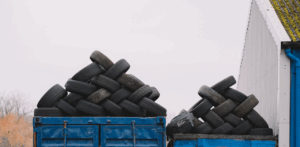 Piles of used car tires are stacked on blue shipping containers outside an industrial building
