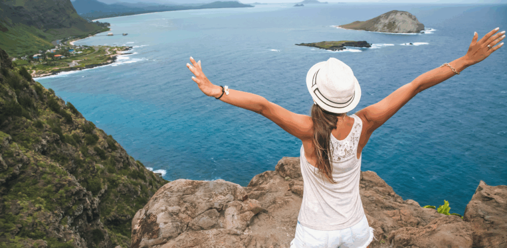 Woman in a white hat standing on a cliff with arms raised, overlooking a stunning tropical ocean view